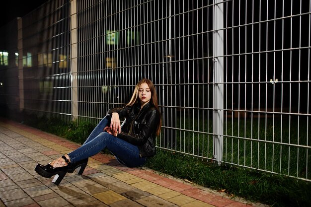 Retrato nocturno de la modelo de niña vestida con jeans y chaqueta de cuero contra la verja de hierro