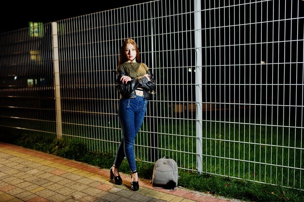 Retrato nocturno de la modelo de niña vestida con jeans y chaqueta de cuero contra la verja de hierro