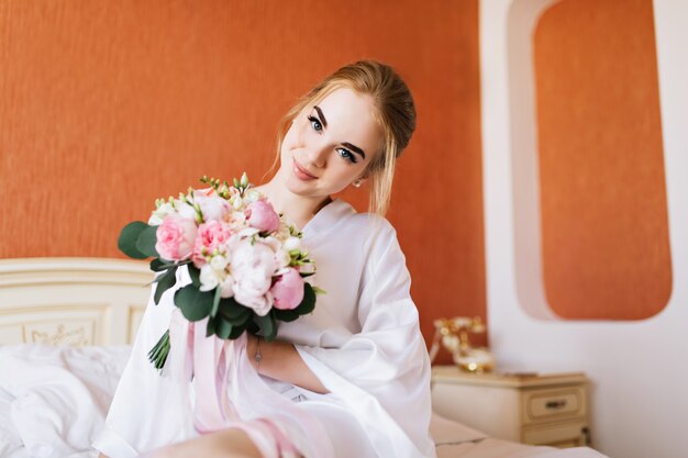 Retrato de novia feliz en bata de baño blanca en la cama por la mañana. Ella sostiene un ramo de flores en las manos