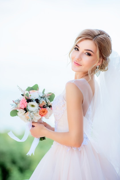 Retrato de una novia impresionante con el pelo rubio que sostiene el ramo de la boda del melocotón en sus brazos