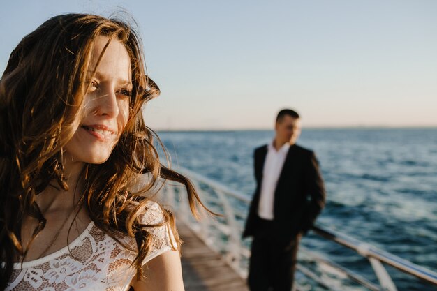 Retrato de una novia morena en primer plano y una silueta de novio en el muelle junto al mar