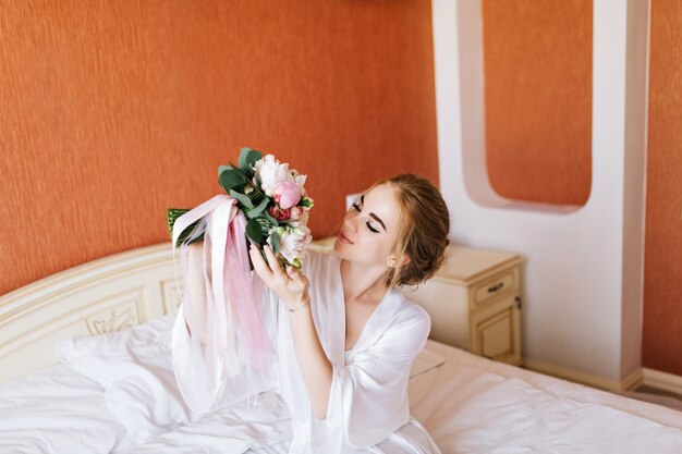 Retrato de novia muy feliz en bata de baño blanca en la cama por la mañana. Ella mira el ramo en las manos y se ve feliz.