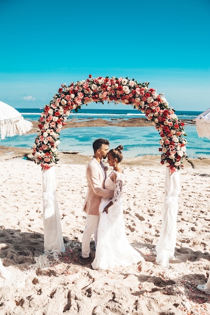 Retrato de la novia y el novio posando junto a la boda arco tropical en la playa detrás del cielo azul y el mar. Pareja de boda