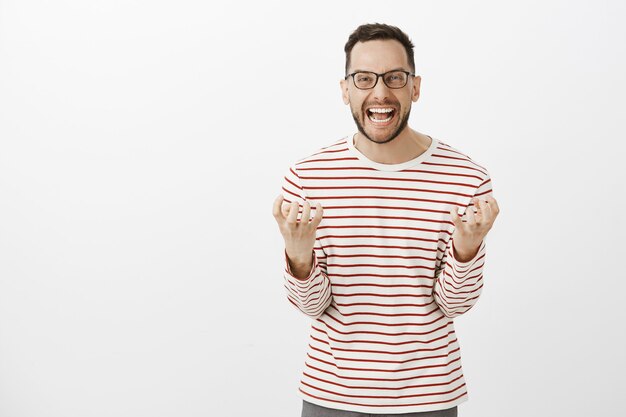 Retrato de un novio adulto europeo deprimido enojado con gafas, gritando en voz alta y apretando los puños, enojado e indignado mientras se pelea con su esposa, queriendo divorciarse sobre una pared gris