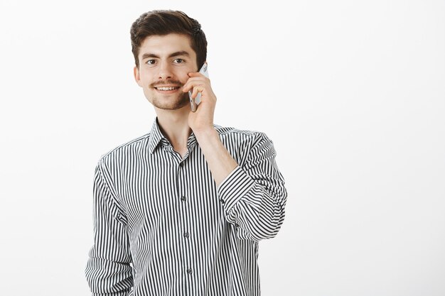 Retrato de novio caucásico despreocupado ordinario con barba y bigote, hablando por teléfono inteligente y sonriendo con alegría, sintiéndose confiado y complacido mientras le pregunta a la chica por una cita sobre una pared gris