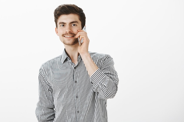 Retrato de novio caucásico despreocupado ordinario con barba y bigote, hablando por teléfono inteligente y sonriendo con alegría, sintiéndose confiado y complacido mientras le pregunta a la chica por una cita sobre una pared gris