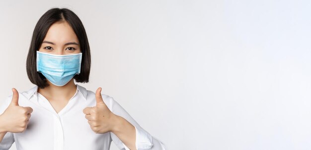 Retrato de una oficinista asiática sonriente con mascarilla médica mostrando los pulgares hacia arriba recomendando algo de pie sobre fondo blanco.