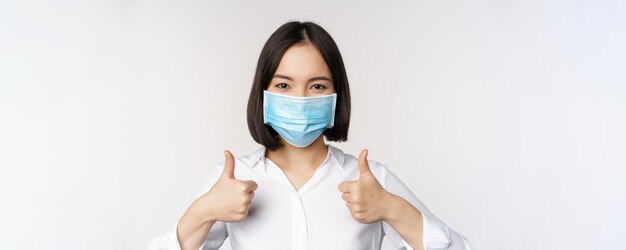 Retrato de una oficinista asiática sonriente con mascarilla médica mostrando los pulgares hacia arriba recomendando algo de pie