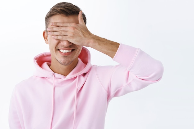 Retrato de optimista joven rubio celebrando un cumpleaños cierra los ojos con la palma y sonriendo anticipando sorpresa, esperando el momento para finalmente ver el presente, la pared blanca