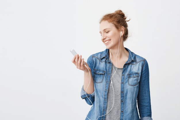 Retrato de optimista positiva jengibre joven mirando la pantalla del teléfono inteligente mientras escucha música o mira videos en los auriculares