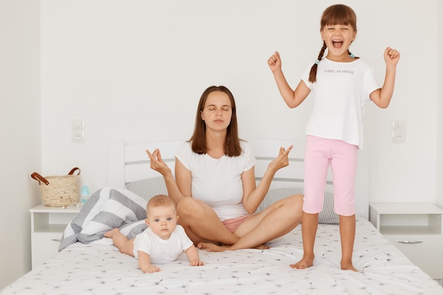 Retrato de otro adulto joven haciendo ejercicios matutinos en pose de yoga mientras sus pequeñas hijas jugando cerca de mamá, niña en edad preescolar de pie con los puños cerrados, niño acostado boca abajo.