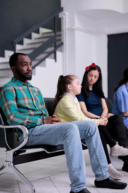 Retrato de un paciente afroamericano que parece cansado esperando en la concurrida sala de recepción del hospital con diversas personas. Hombre aburrido sentado mirando pensativo esperando a que la recepcionista lo llame por su nombre.