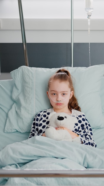 Retrato de paciente hospitalizado niña enferma sosteniendo oso de peluche descansando en la cama durante la co ...