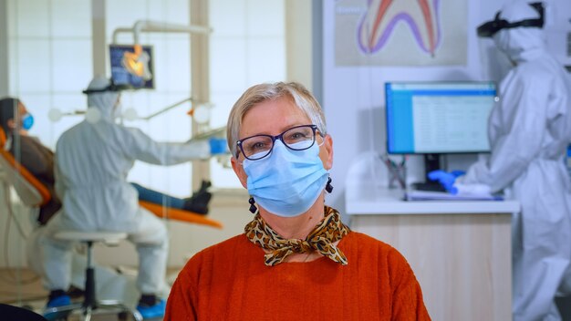 Retrato de paciente jubilado en consultorio dental mirando a cámara con mascarilla sentado en una silla en la clínica de la sala de espera mientras el médico trabaja. Concepto de nueva visita normal al dentista en caso de brote de coronavirus