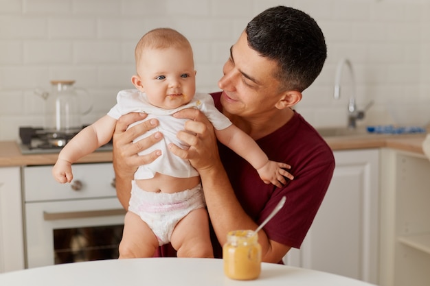 Retrato de padre feliz alimentando a un lindo bebé en camiseta blanca y pañal, sentado en la mesa con comida complementaria, papá con camiseta marrón con linda hija.