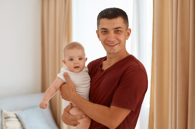 Retrato de padre guapo vistiendo camiseta burdeos con encantadora hija en manos, mirando a cámara, de pie en la acogedora sala de estar con ventana en el fondo.