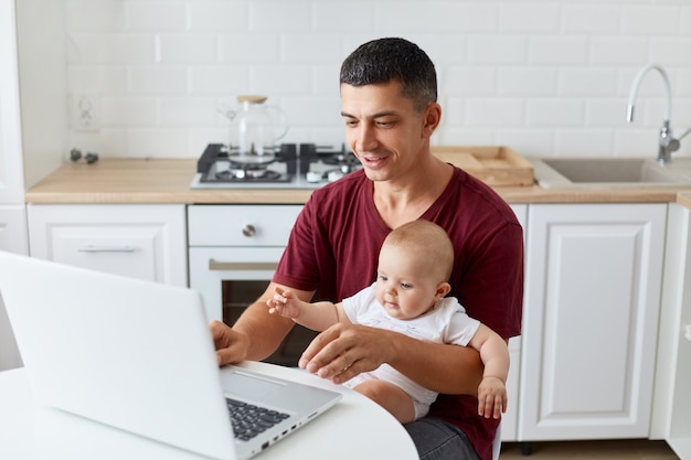 Retrato de padre positivo con camiseta casual granate sentado con el bebé o la niña de rodillas, mirando la computadora portátil con expresión positiva, hombre que trabaja en línea en casa.