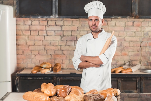 Retrato del panadero masculino que sostiene el rodillo que se coloca detrás de los panes cocidos en la tabla