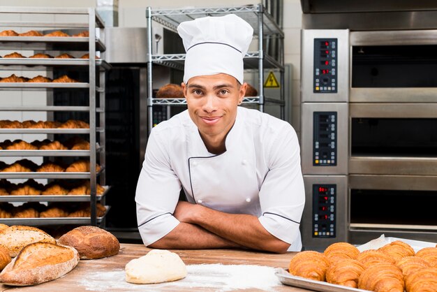 Retrato de un panadero de sexo masculino joven sonriente que se coloca detrás de la tabla con el cruasán y la barra de pan frescos