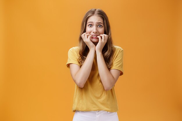 Retrato de pánico nervioso tonto inseguro joven sintiéndose asustado temblando de miedo mordiéndose las uñas manteniendo los dedos en la boca mirando a la cámara asustada sobre fondo naranja.