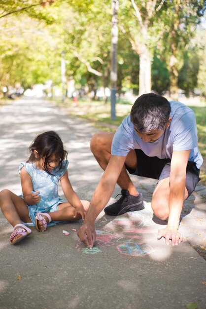 Retrato de papá asiático desarrollando habilidades de dibujo para niños. Hombre feliz y niña sentados en el pavimento en un parque público y dibujando juntos con coloridos crayones. Ocio en verano y concepto de paternidad