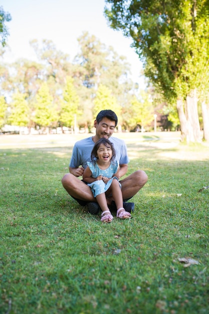 Retrato de papá y su pequeña hija sentada en el césped en el parque. Hombre asiático sosteniendo a una chica agradable de rodillas descansando jugando con alegría en el parque público de verano. Los padres aman, cuidan y el concepto de descanso activo.