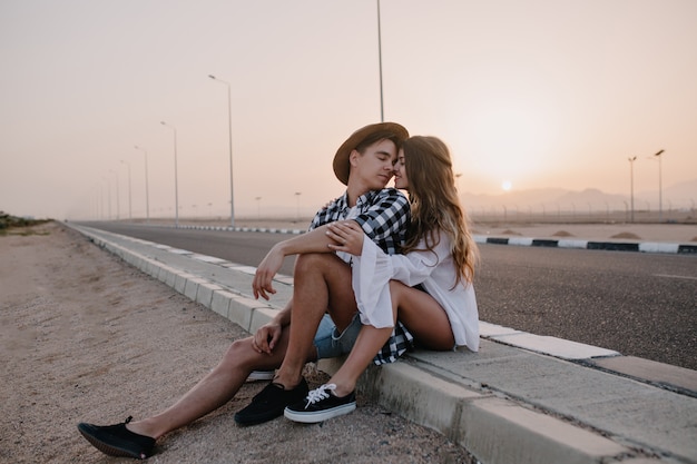 Retrato de pareja amorosa besándose, sentado junto a la autopista después de viajar por la ciudad en fin de semana de verano. Alegre mujer de pelo largo abrazando suavemente a su novio, descansando cerca de la carretera al atardecer