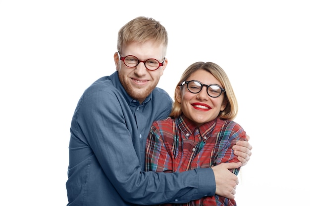 Retrato de una pareja caucásica joven cómica extraordinaria en ropa y gafas excéntricas divirtiéndose: hombre geek con barba de varios días abrazando a su novia atractiva feliz con labios rojos y cabello rubio