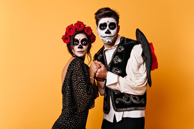 Retrato de pareja de enamorados en pared naranja aislada. Chica y chico en forma de esqueletos se dan la mano con miedo