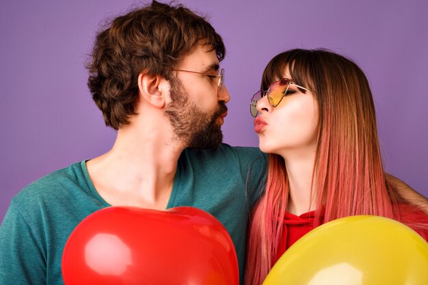 Retrato de pareja feliz hipster mirando el uno al otro y tratando de besarse, sosteniendo globos, ropa casual de moda brillante y gafas, humor romántico de cerca