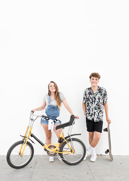 Retrato de una pareja feliz de pie con bicicleta y patineta delante de la pared