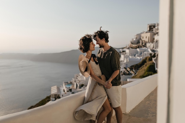 Retrato de pareja en el fondo de la ciudad griega Mujer morena con vestido beige y hombre con traje elegante se miran con amor en el fondo del mar