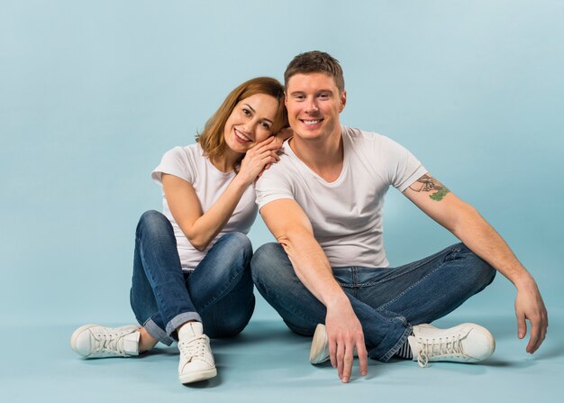 Retrato de una pareja joven amorosa sonriente sentada en el piso contra el fondo azul