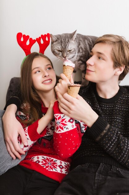 Retrato de una pareja joven y bonita sentada en un sofá en casa con helado en las manos y un buen gato cerca de ese helado que prueba