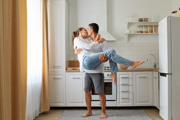 Retrato de pareja joven en una cocina acogedora, el chico y la chica se besan y abrazan, el hombre sostiene a una mujer en sus brazos, pasando tiempo juntos, luna de miel, sentimientos románticos.