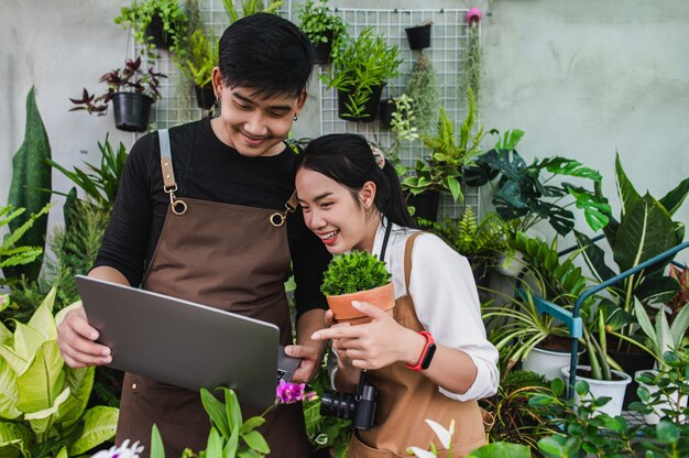 Retrato de pareja joven jardinero asiático con delantal utiliza equipo de jardín y computadora portátil para investigar y cuidar las plantas de la casa en invernadero