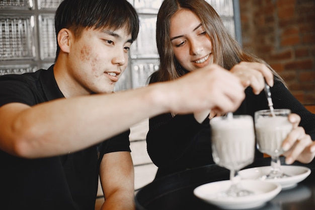 Retrato de pareja joven sentada en el restaurante con capuchino en las manos. Chica caucásica con cabello oscuro y chico asiático con cabello oscuro sentados juntos. Niño y niña con camisetas negras.