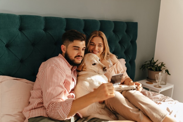 Retrato de pareja joven y su doglying en cama de color esmeralda. Marido y mujer miran una foto memorable con una sonrisa.