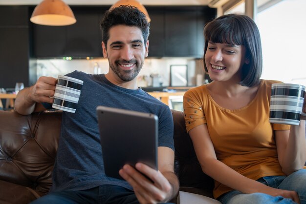 Retrato de pareja joven en una videollamada con tableta digital mientras está sentado en el sofá en casa. Quédate en casa. Nuevo concepto de estilo de vida normal.