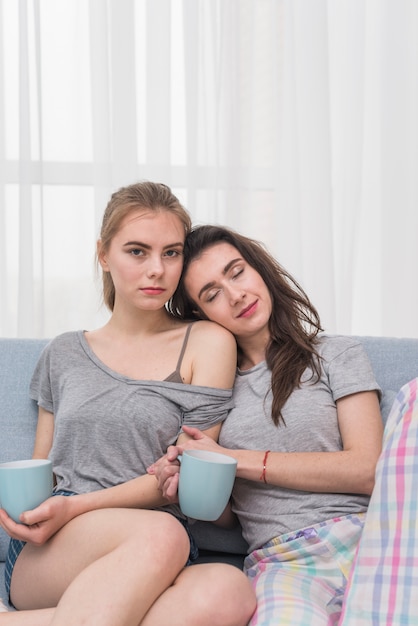 Retrato de una pareja de lesbianas jóvenes sentados en el sofá sosteniendo una taza de café azul