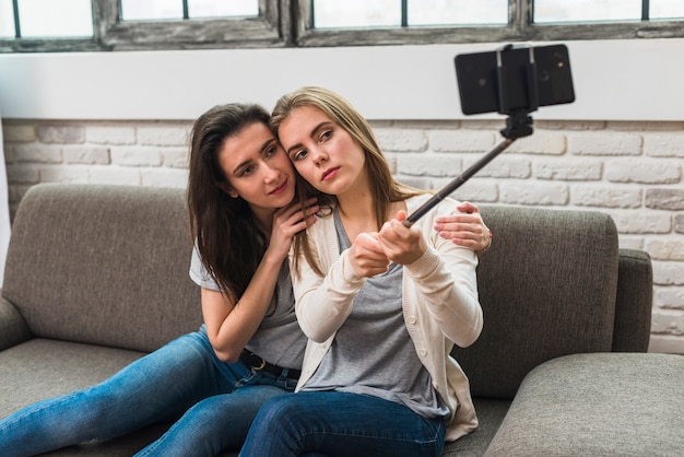 Foto gratuita retrato de una pareja de lesbianas jóvenes sentados en el sofá tomando selfie en el teléfono móvil