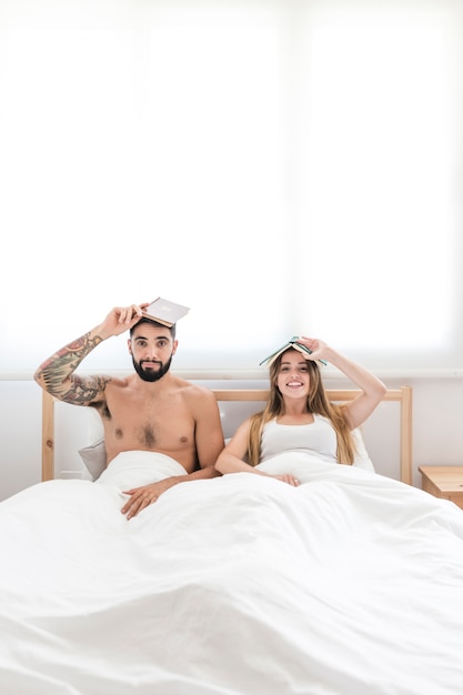 Retrato de pareja sentada en la cama sosteniendo un libro sobre su cabeza