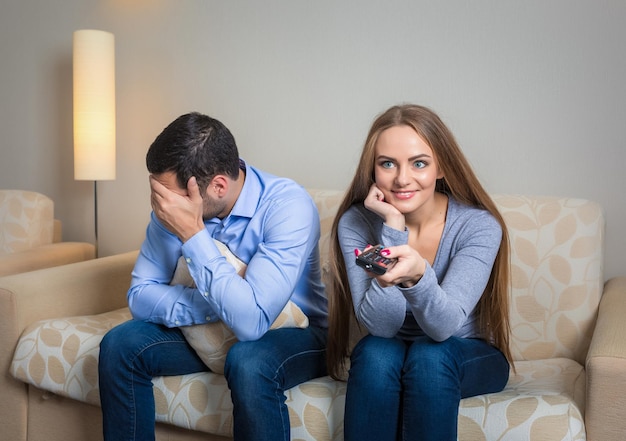 Retrato de una pareja sentada en un sofá viendo la televisión. Imagen de mujer con control remoto en manos y hombres molestos