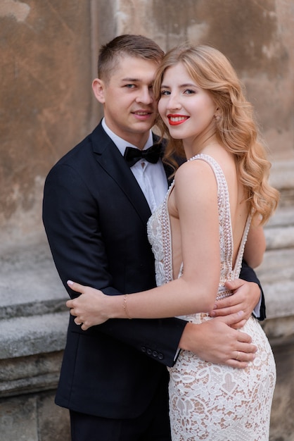 Retrato de pareja sonriente en elegante vestido y esmoquin negro cerca de muro de piedra