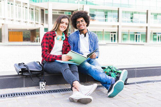 Retrato de los pares étnicos multi jovenes que se sientan delante del edificio de la universidad que estudian juntos