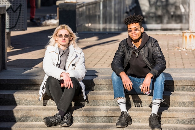 Retrato de los pares interraciales jovenes elegantes que se sientan en escalera