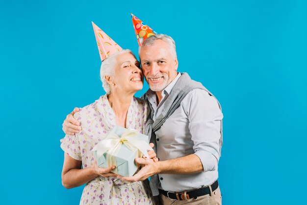 Retrato de los pares mayores felices que sostienen el regalo de cumpleaños en fondo azul