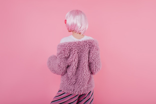 Retrato de la parte posterior del modelo femenino de moda lleva chaqueta de piel. Agraciada mujer caucásica en corto periwig brillante mirando pared rosa