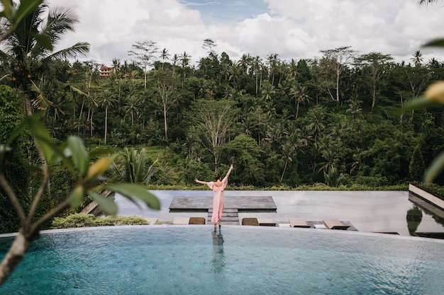 Retrato de la parte posterior del modelo femenino en vestido rosa mirando la selva tropical. Tiro al aire libre de agraciada mujer bailando junto a la piscina.