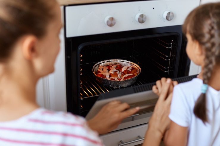Foto gratuito retrato de la parte trasera de una mujer joven que pone croissant en un plato para hornear del horno, su hija de pie cerca y mirando dentro de la estufa de gas, quiere tener un sabor delicioso lo antes posible.
